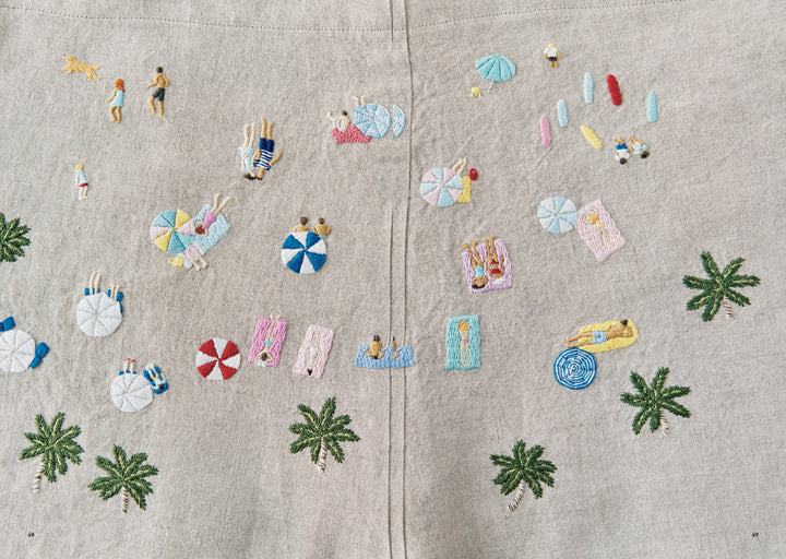 Close-up of the Juno's Nature Embroidery Notebook by Schiffer Publishing, Ltd., featuring playful beach-themed and nature embroidery patterns with people, palm trees, umbrellas, and accessories on beige fabric.