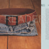 A copy of "A Guide to Making Leather Belts with 12 Complete Projects" by Schiffer Publishing, Ltd. rests beside a brown leather belt atop folded blue jeans, all placed on a wooden surface. A light blue title page with decorations appears to the right.