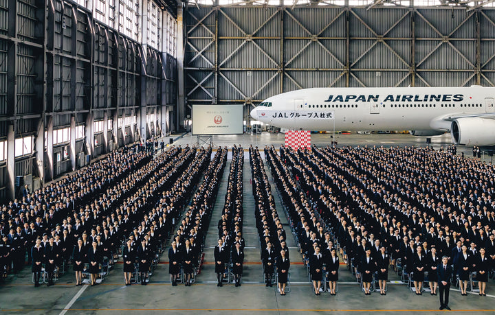 Hundreds in dark suits line up inside a hangar with a Japan Airlines plane and a screen behind—a striking image from Japan by Schiffer Publishing, Ltd., perfect for any travel photography collection.
