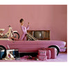 A woman in retro attire poses with a pink bicycle and the Mini Honda by Schiffer Publishing, Ltd. on a pink convertible, surrounded by vintage luggage and household items, against a vibrant pink background.
