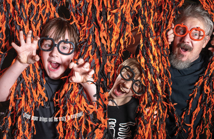Three people wearing black shirts and playful Halloween glasses pose with clawed hands among orange and black streamers, showing off creative Halloween decor inspired by Schiffer Publishing, Ltd.'s "How to Haunt Your House Halloween Craft Fun.