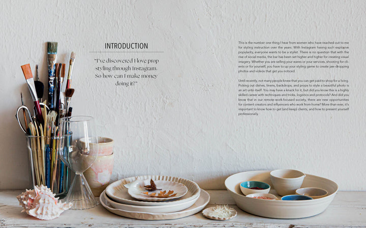 A tabletop with paintbrushes, glassware, seashells, and ceramic dishes is set before a white textured wall featuring introductory text from "Styling Beyond Instagram" by Schiffer Publishing, Ltd. on making money in prop styling.